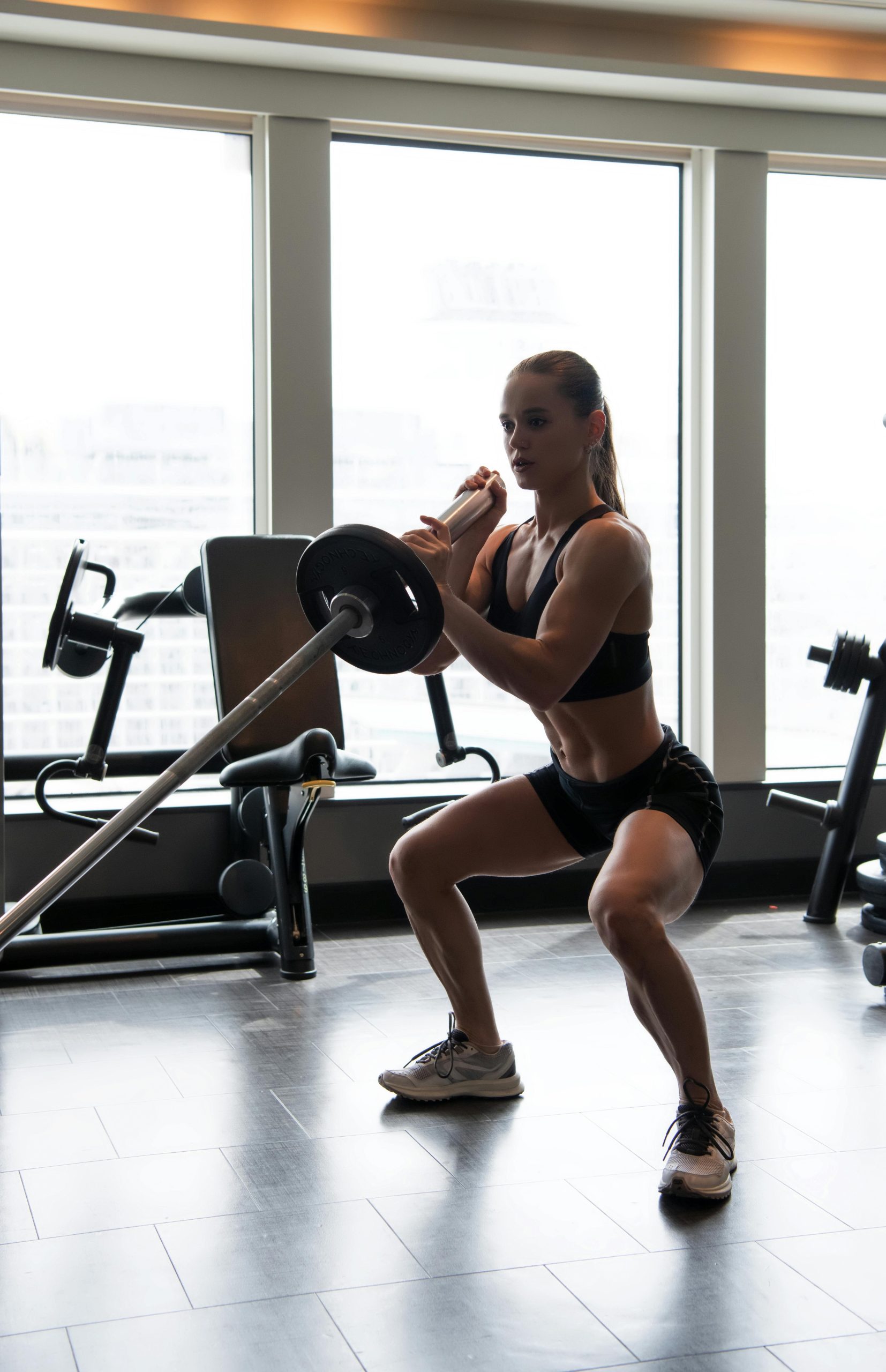 A dedicated female athlete executing a barbell squat in a modern gym setting during the day.
